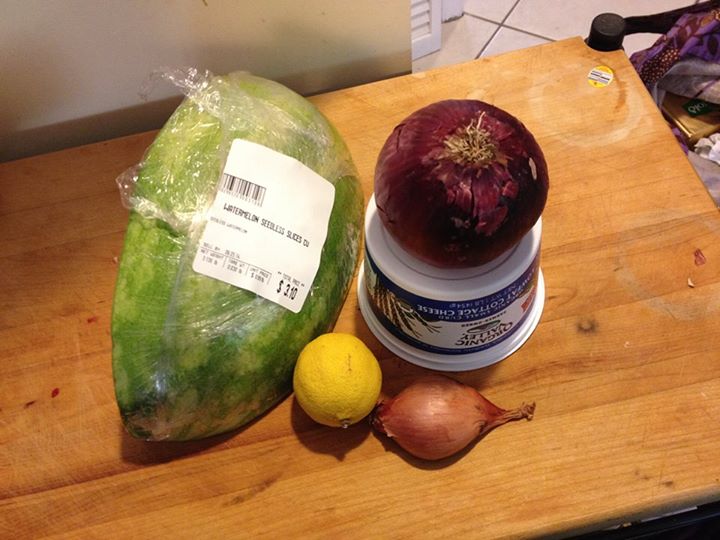 A quarter of a smallish watermelon, a red onion, a lemon, and a lobe of shallot sitting pertly on a cutting board.